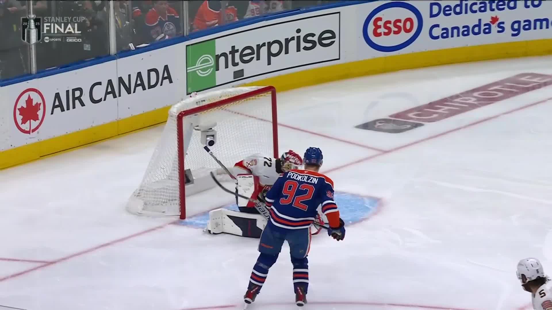 The goalie, in red, is crouched low in front of the net, while a player in blue and orange skates toward him. Number 92 is on the back of the player's jersey.
