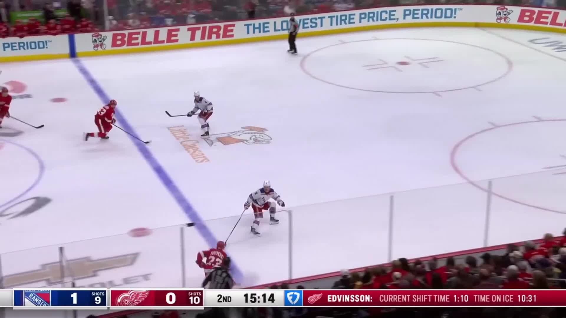 A player in red skates toward the puck, stick extended, as two opponents in white and gray try to intercept. The scoreboard shows the Rangers leading the Red Wings in the second period of the NHL game.
