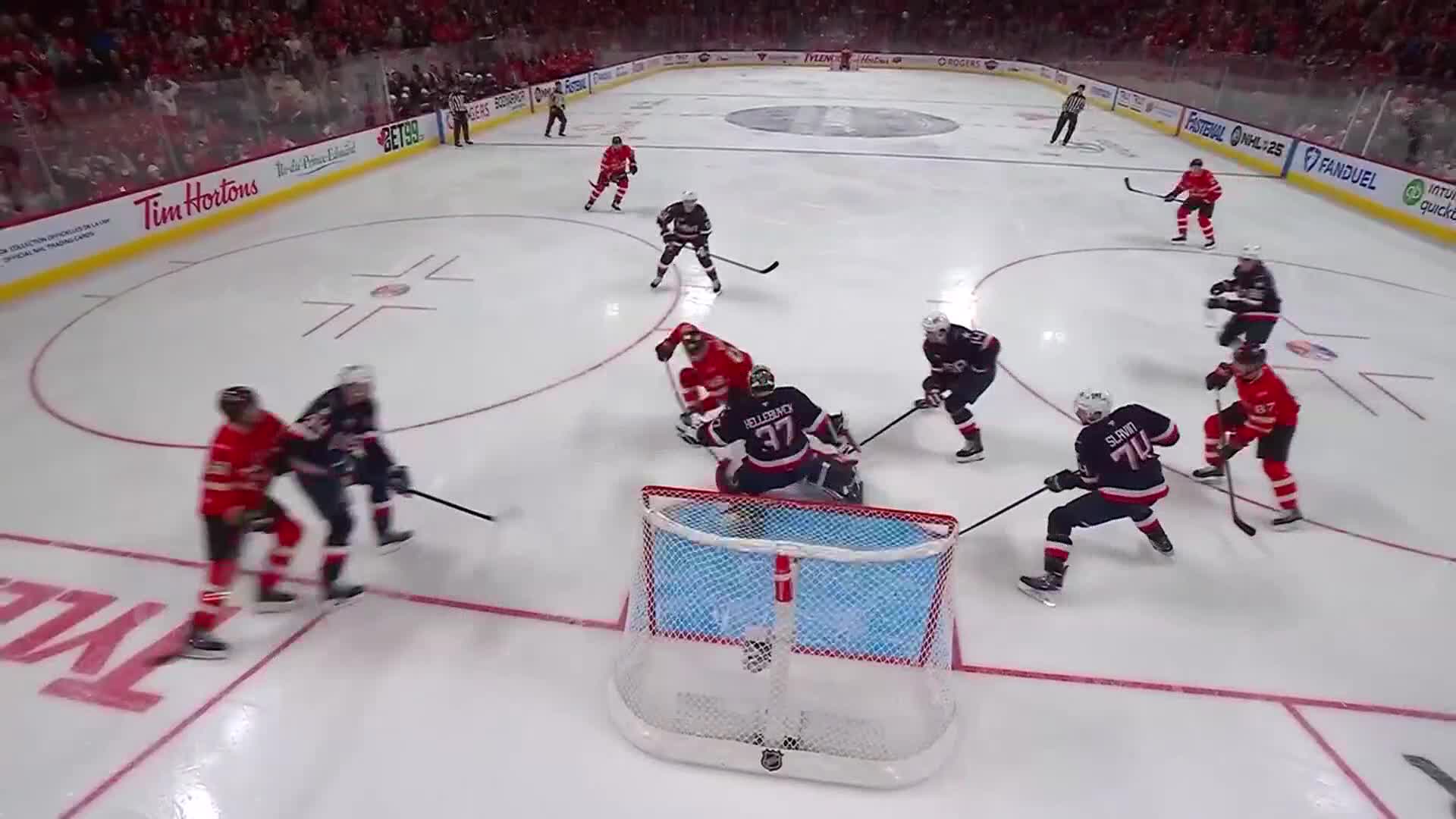 The goalie, in black, is braced in front of the net as a red-jerseyed player tries to get past him. A player in a dark jersey with the number 74 is skating toward the net, stick raised.
