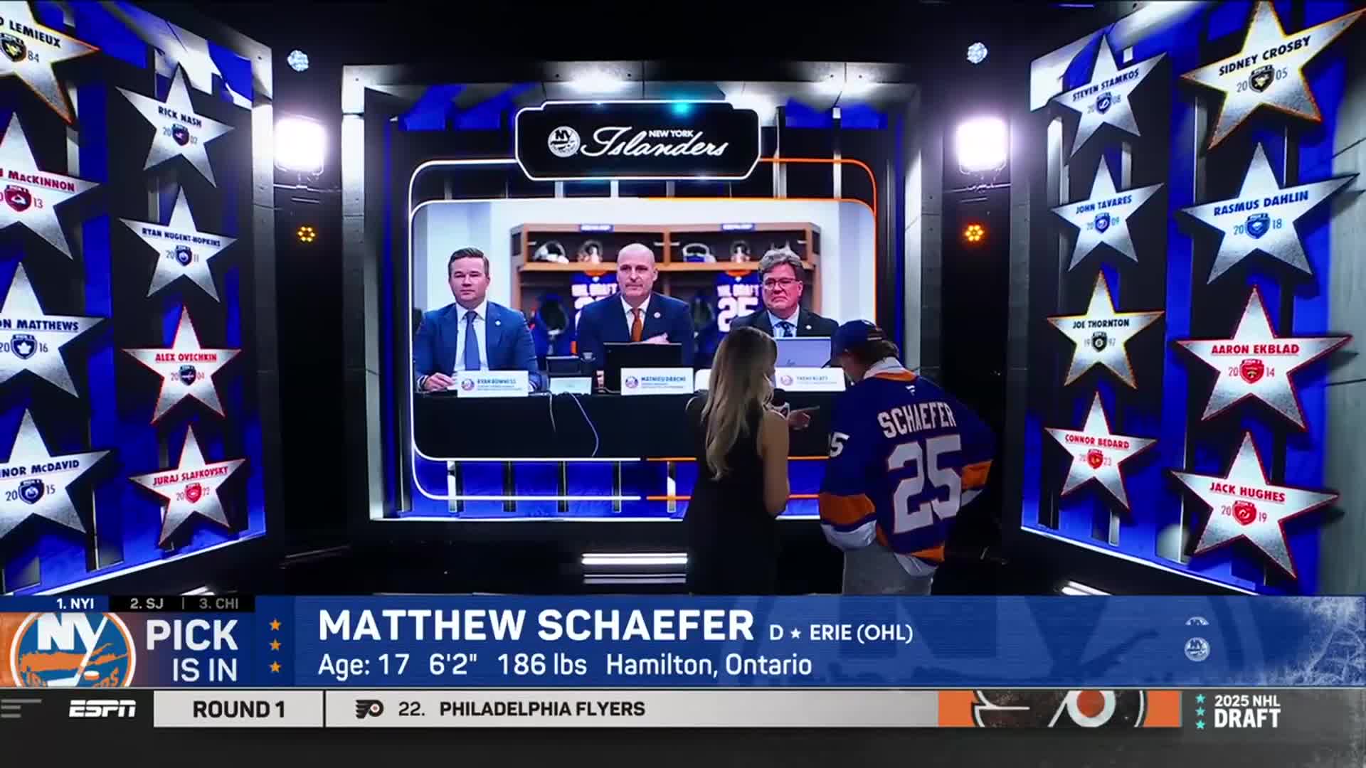 A young man in a New York Islanders jersey stands next to a woman, both facing a screen showing the NHL Network broadcast. The screen displays the team's selection of Matthew Schaefer, along with the team's front office.
