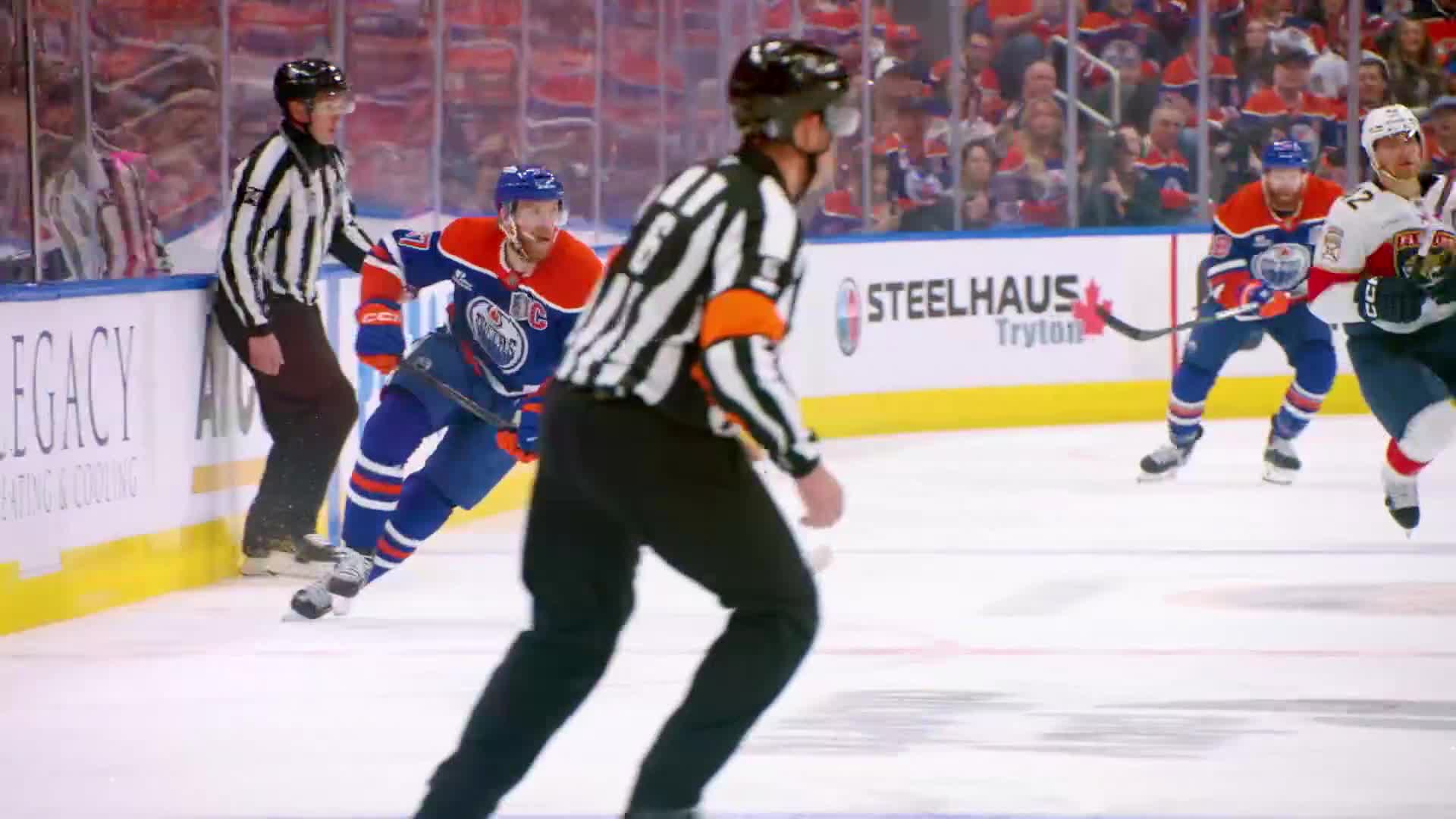 The referee in black and white stripes skates forward, focused on the play. An Oilers player in blue and orange follows the puck up the ice, with another player from the opposing team close behind.
