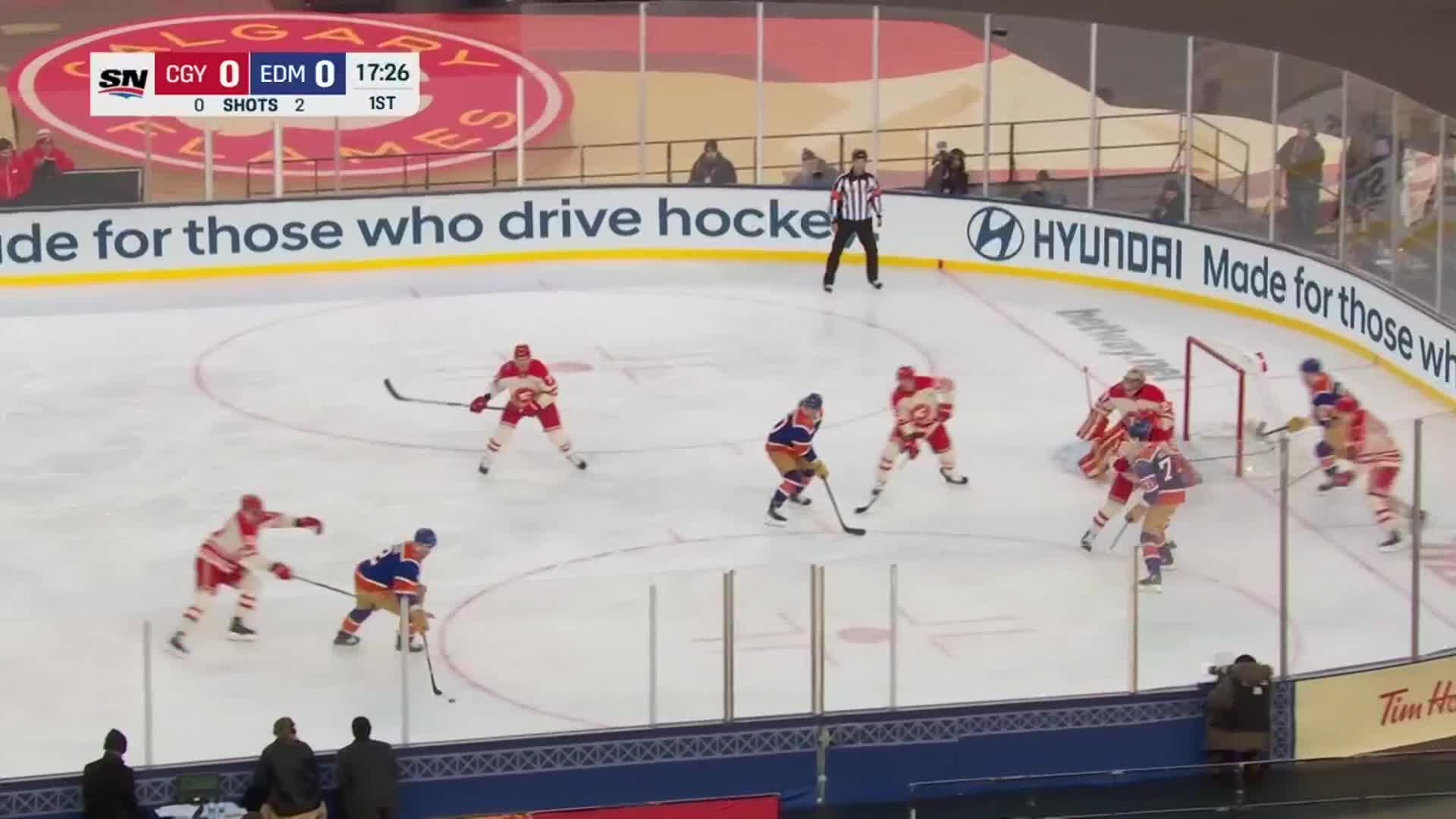 An Edmonton player in blue and orange skates with the puck, pursued by Calgary Flames in red. The puck carrier is near the net, with a goalie in red ready to defend.
