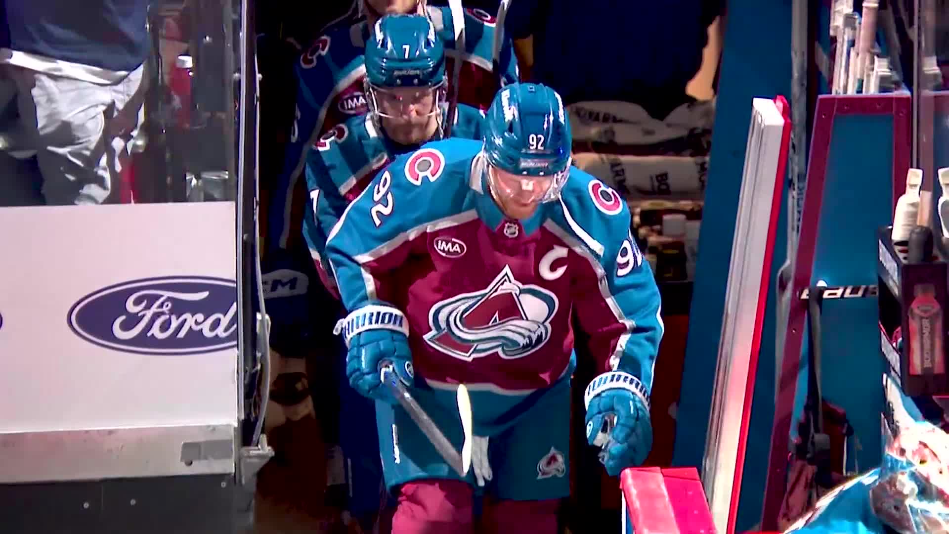 The Colorado Avalanche players, in their teal and burgundy jerseys, are heading onto the ice. Number 92, with the "C" on his chest, leads the way, stick in hand.
