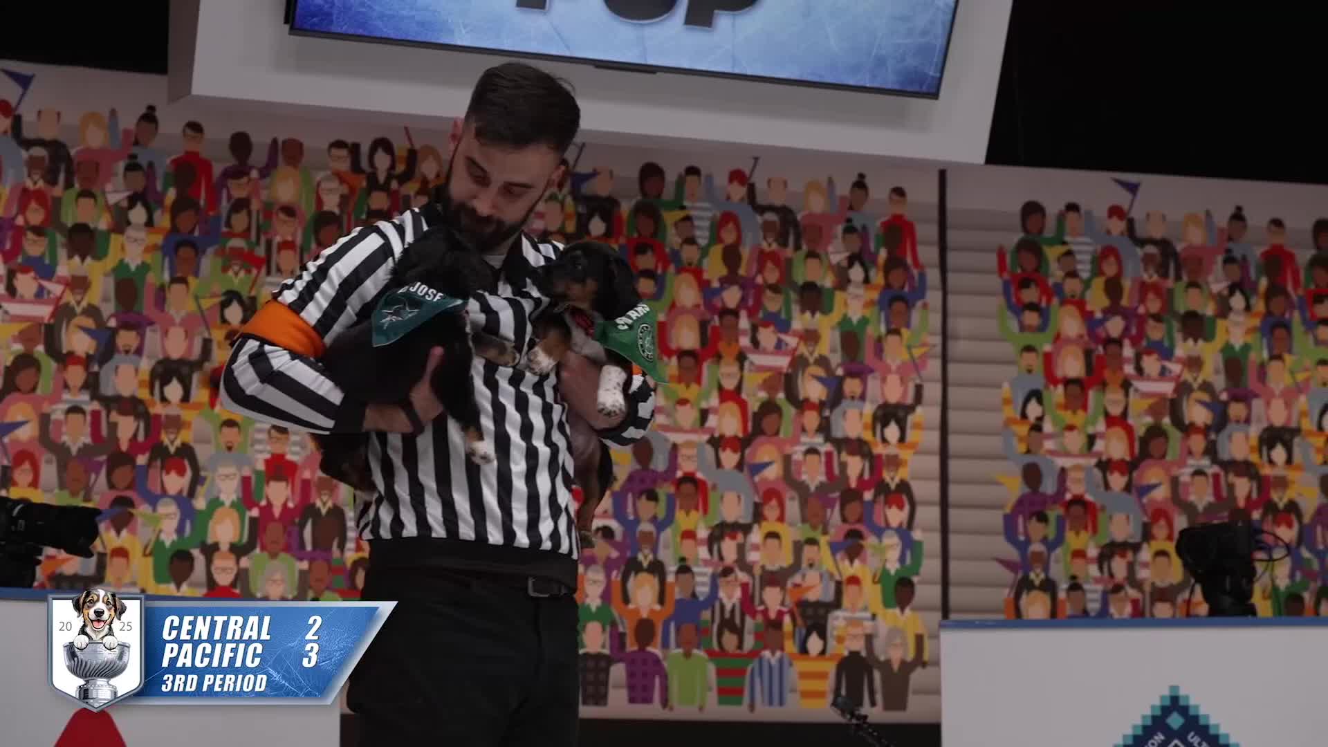 An official, in a striped shirt, cradles two small dogs, each wearing a bandana. The scoreboard shows the Central and Pacific teams playing in the third period.
