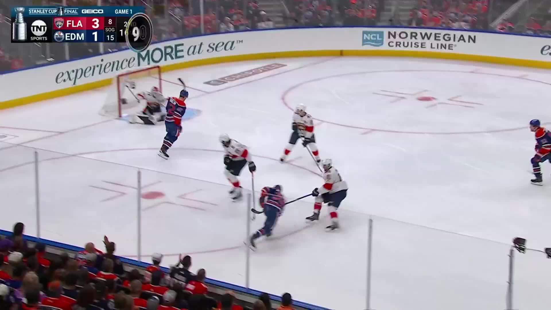 An Edmonton Oilers player, in blue and orange, skates toward the net, stick raised. The Florida Panthers defense, in white and red, attempts to block his shot in this NHL game.
