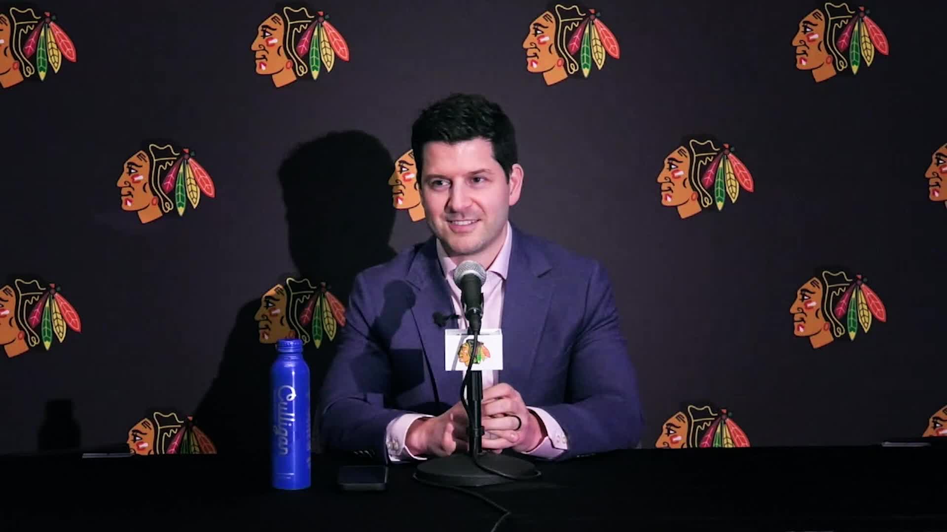 A man in a suit sits at a table, speaking into a microphone with the Blackhawks logo on it. Behind him, the team's logo repeats across a black backdrop.
