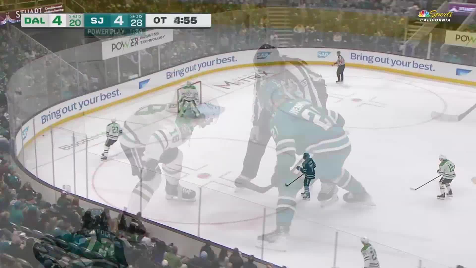 A player in a green jersey skates with the puck toward the net. Another player in a white jersey prepares to receive a pass near the blue line.