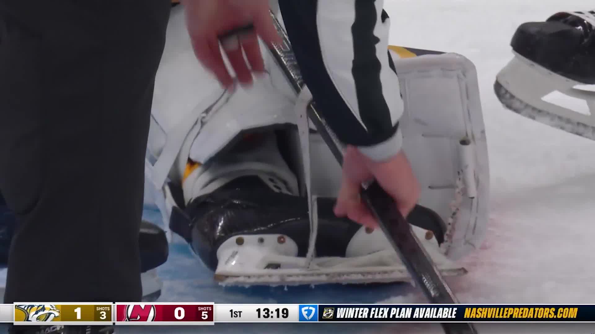 A referee adjusts his grip on the hockey stick, his striped sleeve brushing against the goalie's pads. The scoreboard in the bottom corner indicates the Predators are up 1-0 in the first period.