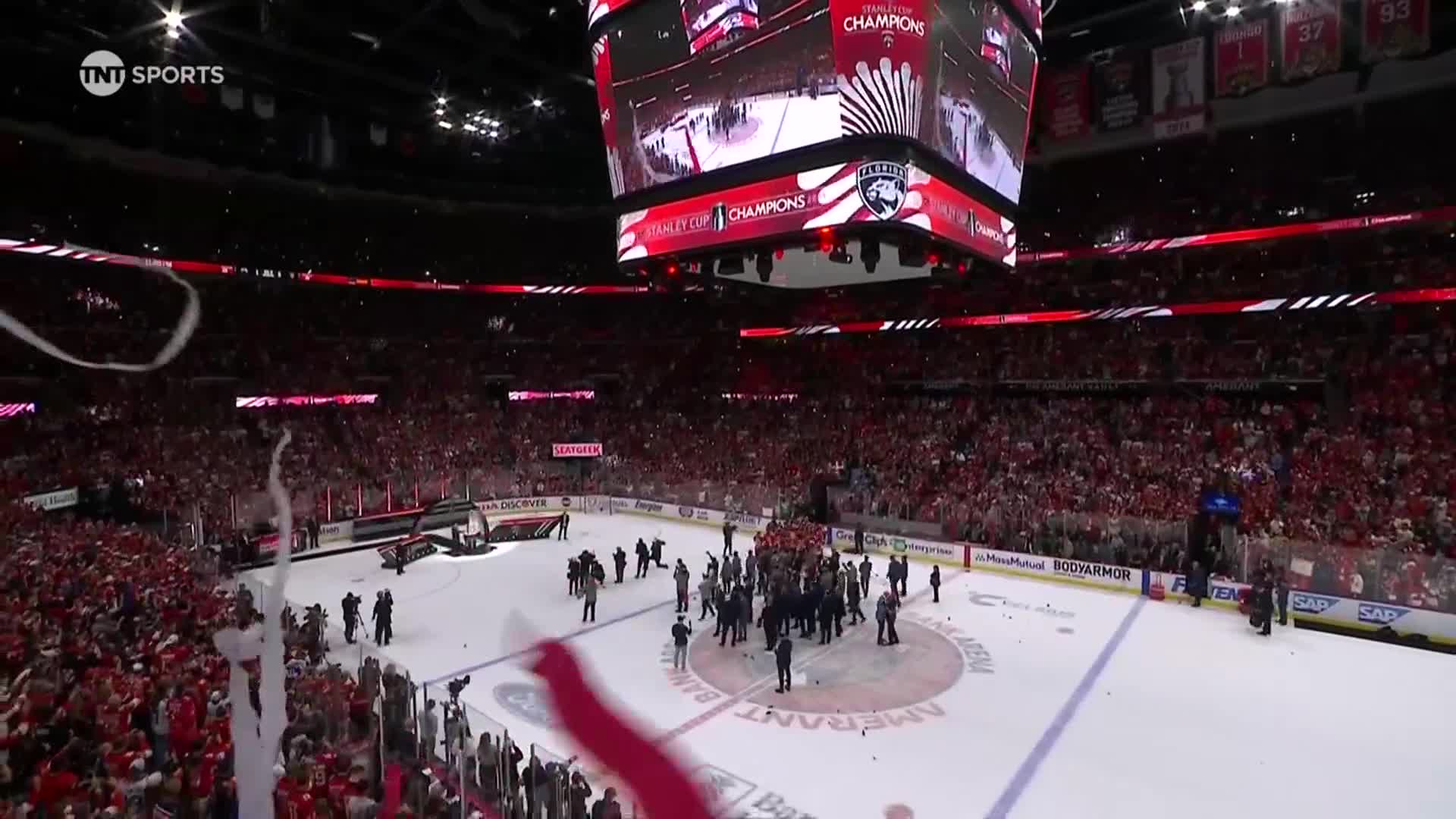 Confetti rains down on the ice as players gather around the Stanley Cup. The scoreboard above flashes "Stanley Cup Champions."