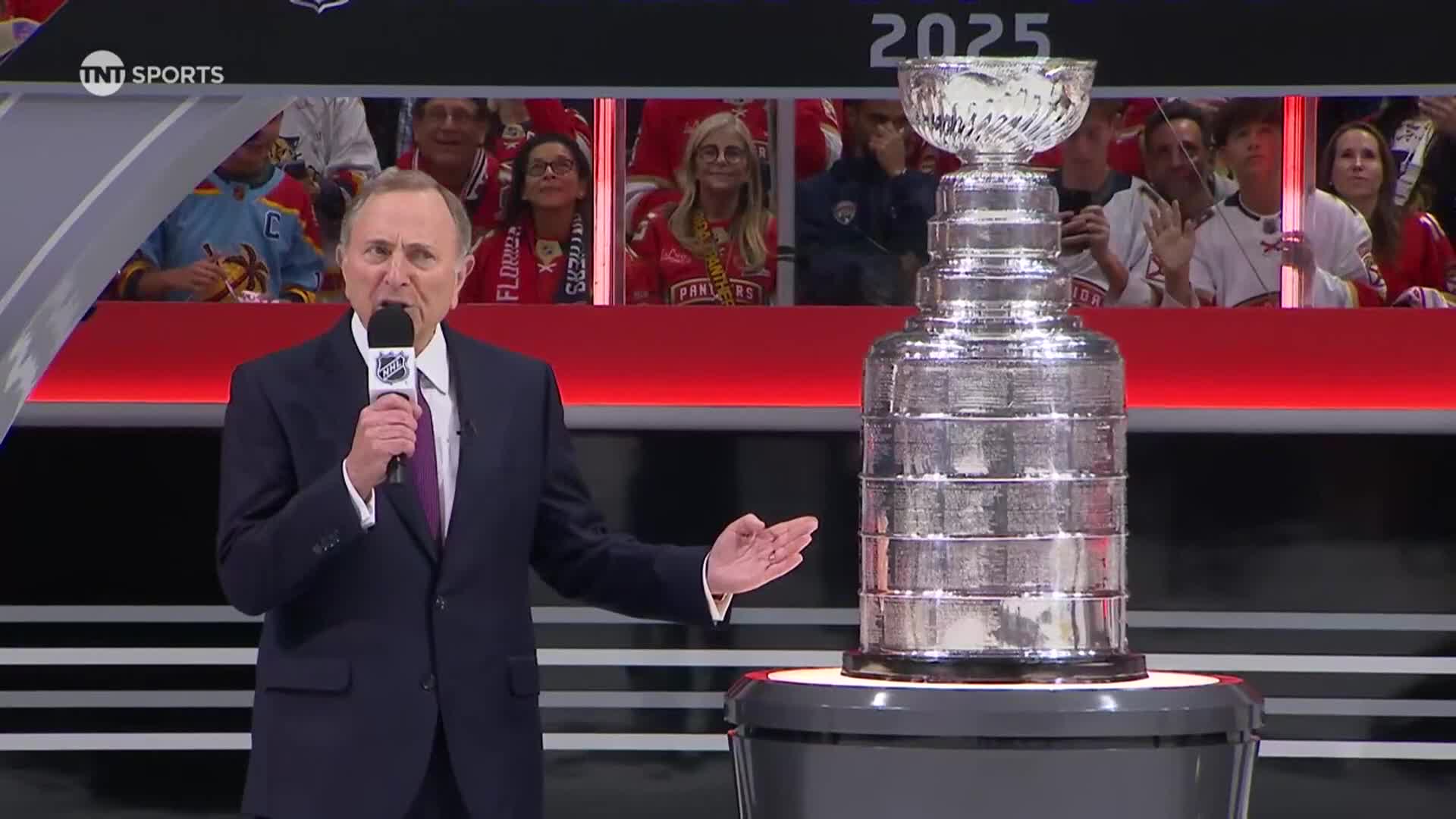 A man in a suit speaks into a microphone, gesturing toward the Stanley Cup. The trophy gleams under the lights, its many rings etched with names.
