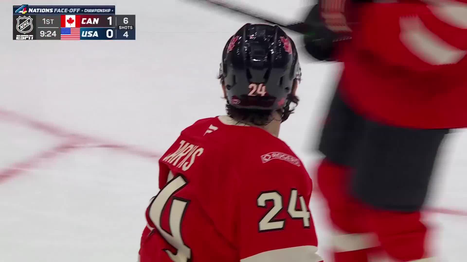 A player in a red jersey with the number 24 on his back skates past. The scoreboard shows Canada leading the USA 1-0 in the first period.