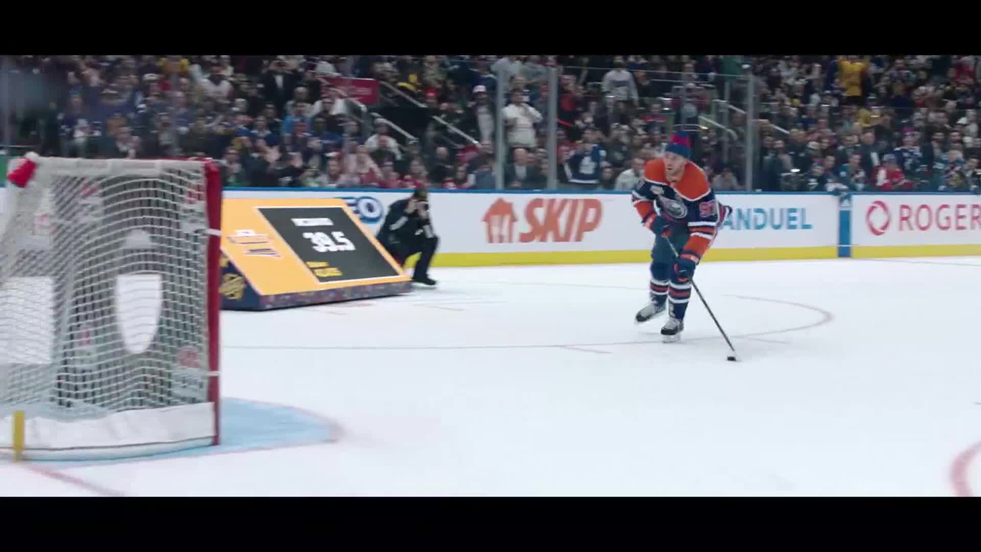 A hockey player in an orange and blue jersey skates toward the net, stick on the ice. A referee crouches near a digital scoreboard displaying "39.5."
