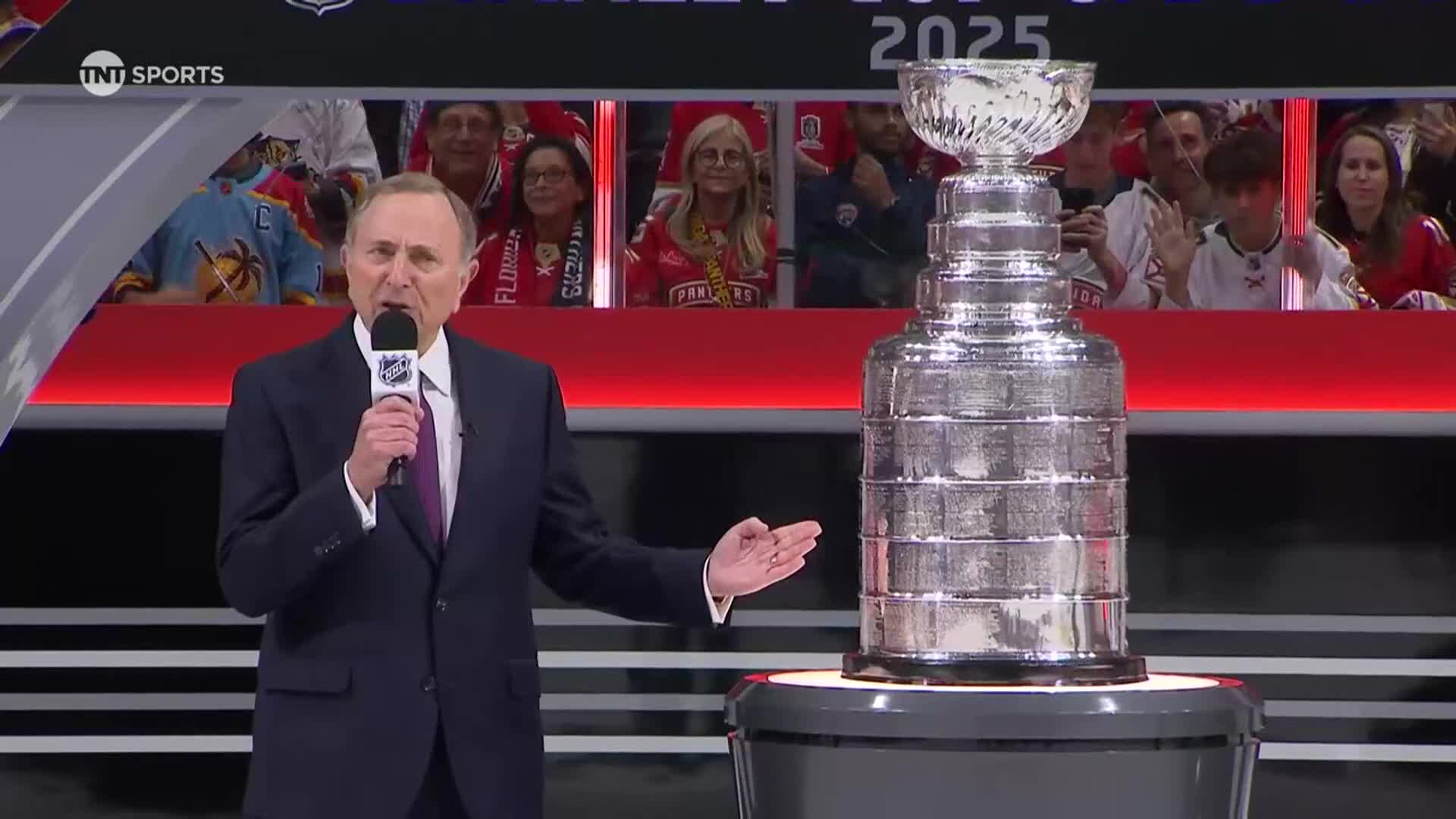 A man in a suit speaks into a microphone beside the Stanley Cup. Spectators in the background wear team jerseys.