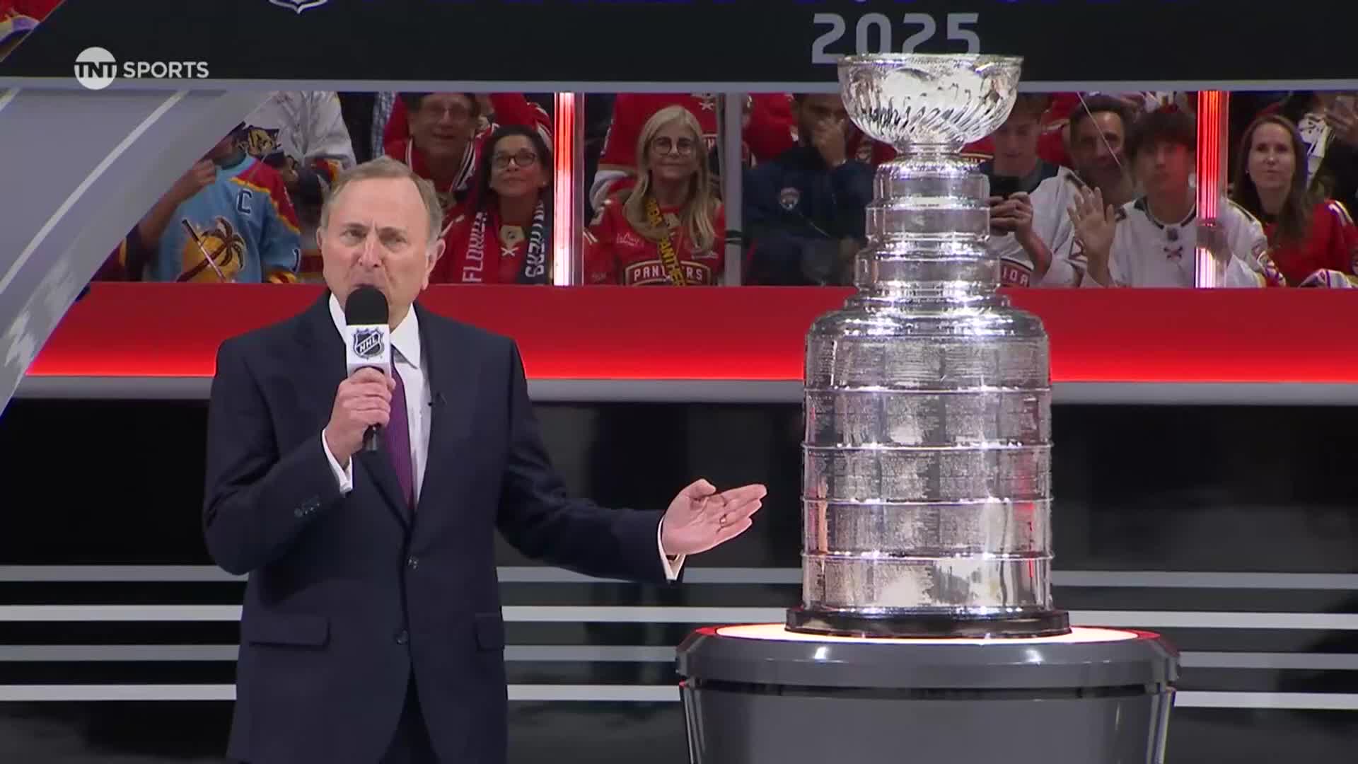 A man in a suit speaks into a microphone next to the Stanley Cup. Behind him, fans in red jerseys watch from the stands.