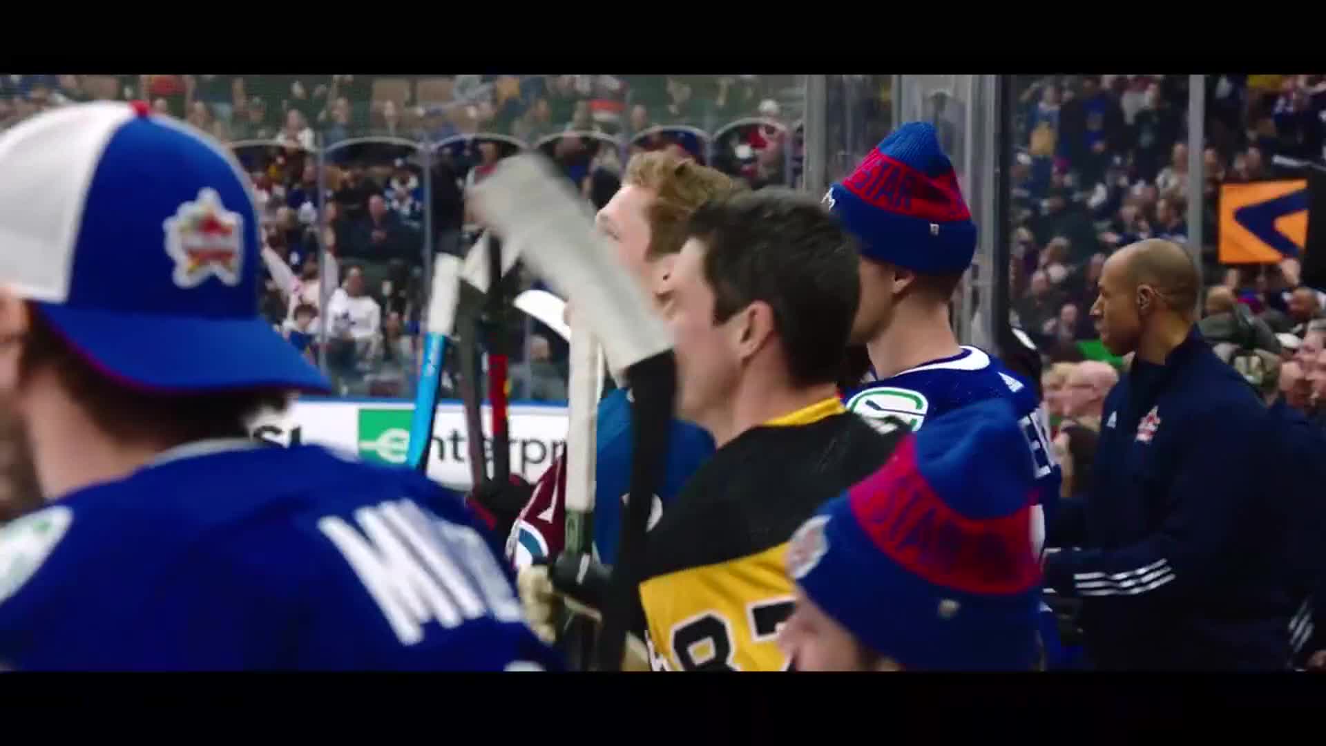 Players in blue and yellow jerseys stand by the boards, some wearing knit hats. A man in a dark jacket with red and white stripes walks past them.