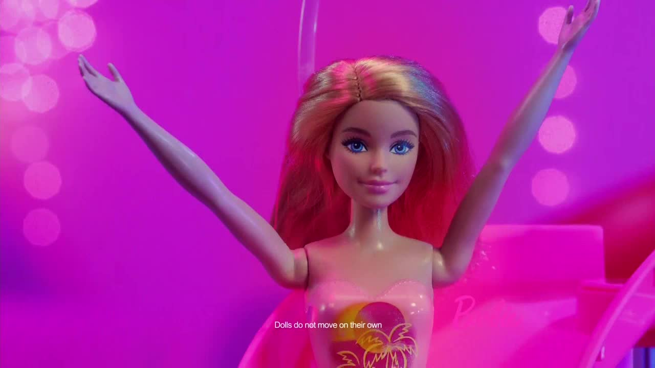 A Barbie doll, with her arms raised, stands in front of a bright pink background. The doll's hair is a mix of blonde and pink, and she's wearing a pink top.
