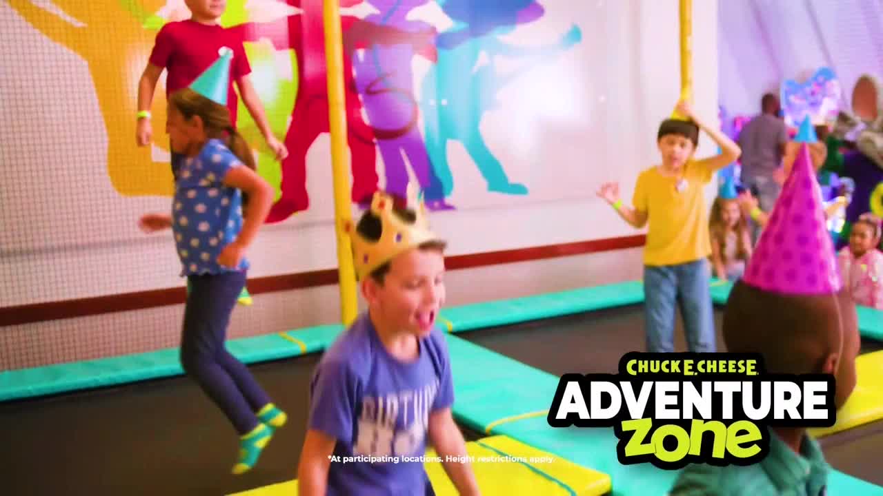 Kids in party hats bounce on trampolines, their faces lit up with excitement. A boy in a crown yells as he leaps, while others run and jump with glee.
