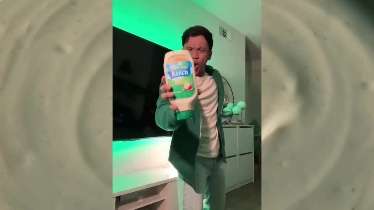 A man in a green jacket holds up a bottle of ranch dressing, yelling towards the camera. The background is a living room with a green-lit television and a white cabinet.
