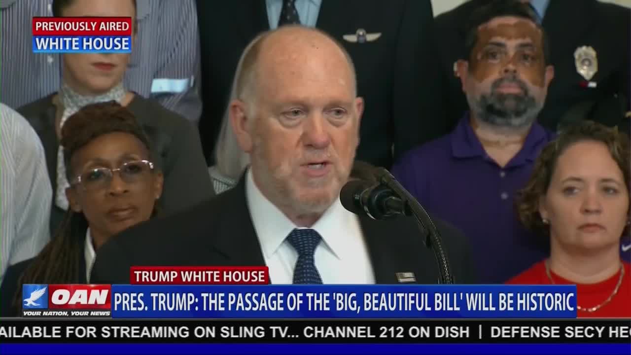A man in a dark suit stands at a podium, speaking into a microphone. The One America News Network logo is visible at the bottom of the screen, along with a headline about President Trump.

