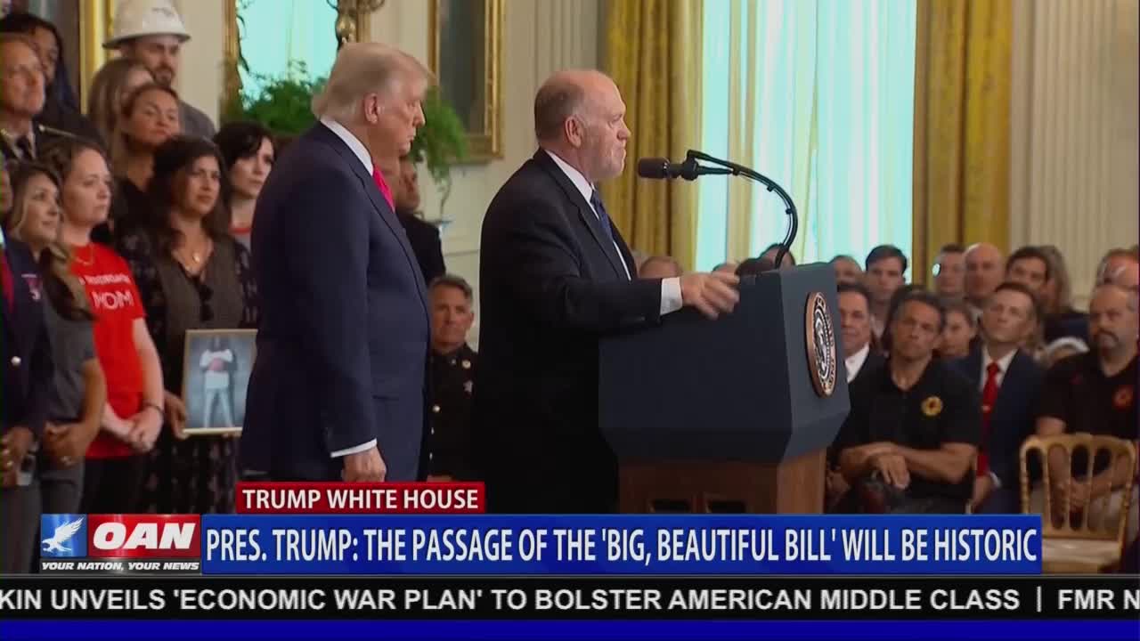 A man in a suit speaks at a podium, while Donald Trump stands beside him, looking on. A crowd of people, some holding framed photos, watches the event, which is covered by One America News Network.
