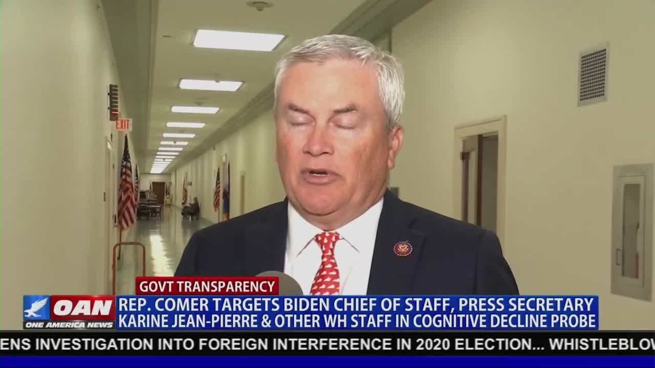 A man in a suit and red tie speaks directly into a microphone, framed by a hallway in the United States. The lower third of the screen, from One America News Network, announces a probe targeting White House staff.
