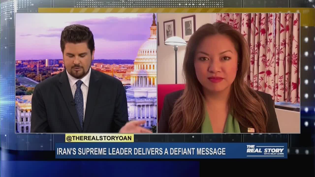 A man in a suit, likely in a studio in the United States, is looking down and talking, while a woman on the right side of the screen appears to be in a remote interview. The lower third of the screen displays a headline about Iran, and the show's name, "The Real Story" is visible.
