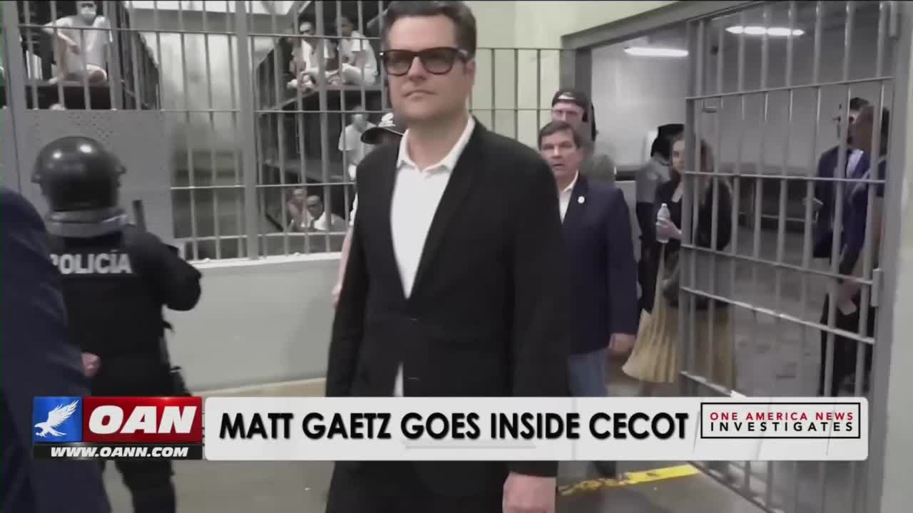 Matt Gaetz, wearing sunglasses and a suit, walks toward the camera through a doorway with bars. Behind him, a group of people follows, while a police officer in riot gear stands to the left.
