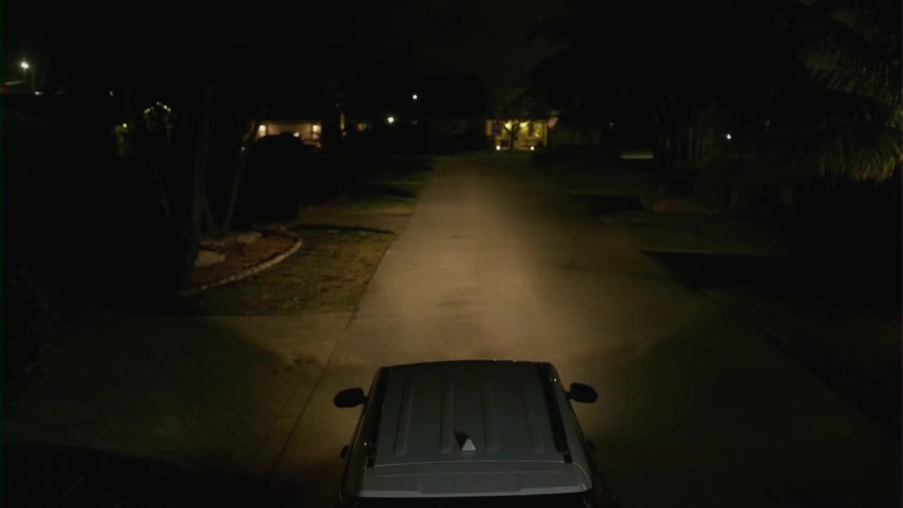 A vehicle's headlights illuminate a dark residential street. The car is moving forward, its roof visible in the foreground.
