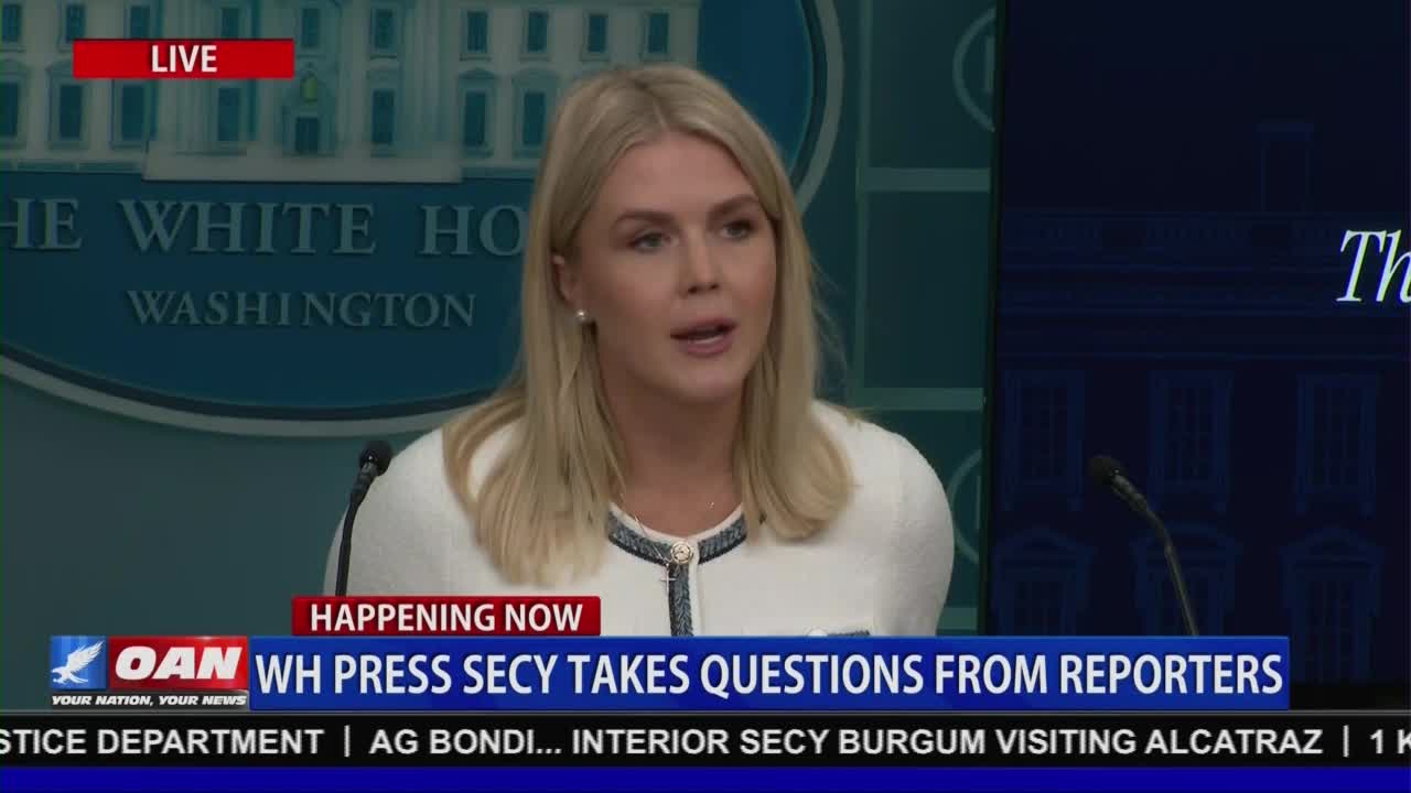 A woman with blonde hair, wearing a white jacket, speaks into a microphone at a podium. The live broadcast on One America News Network is reporting on the White House Press Secretary taking questions from reporters.
