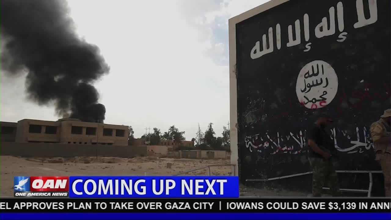 A building is engulfed in thick, black smoke, billowing into the sky. On the right, a large sign displays Arabic script and an emblem, while a news graphic from One America News Network flashes at the bottom.

