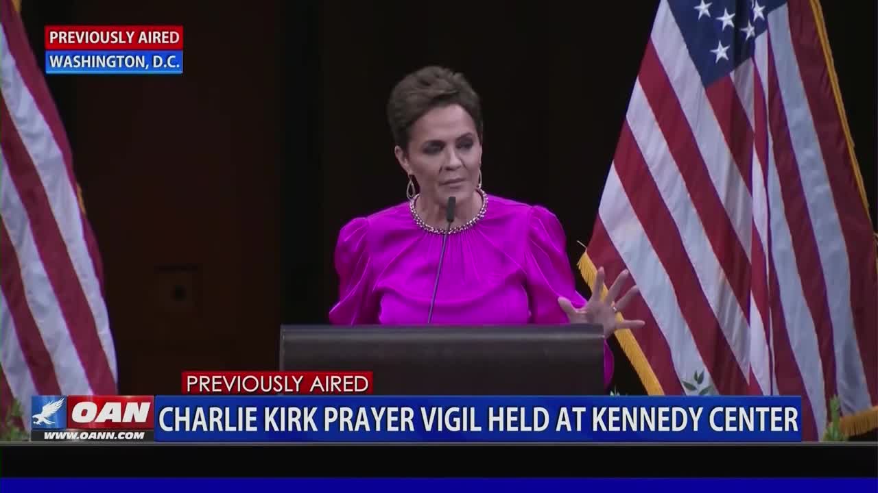 A woman in a bright pink dress stands behind a podium, gesturing with her hands. The backdrop includes American flags and a lower-third graphic from One America News Network.
