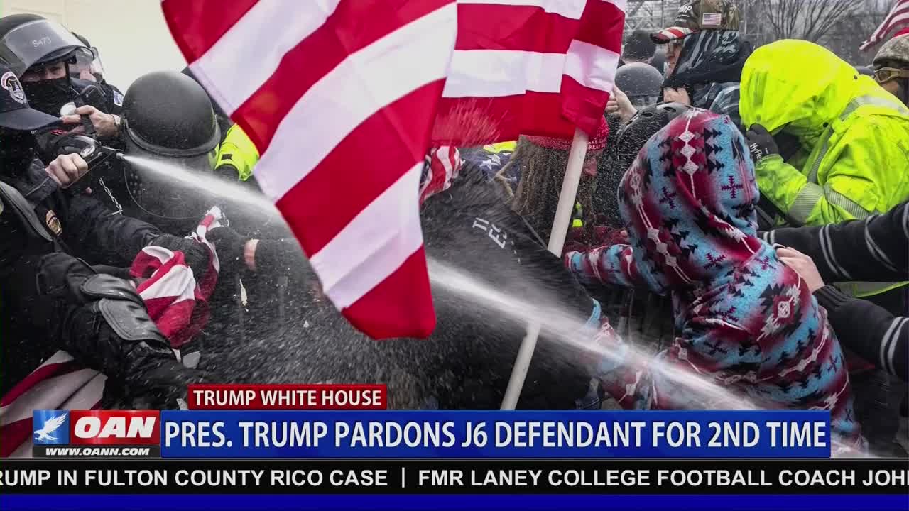 A high-pressure water stream blasts toward a crowd, obscuring the faces of those holding an American flag. The scene unfolds as a news graphic from One America News Network reports on a Trump White House pardon.
