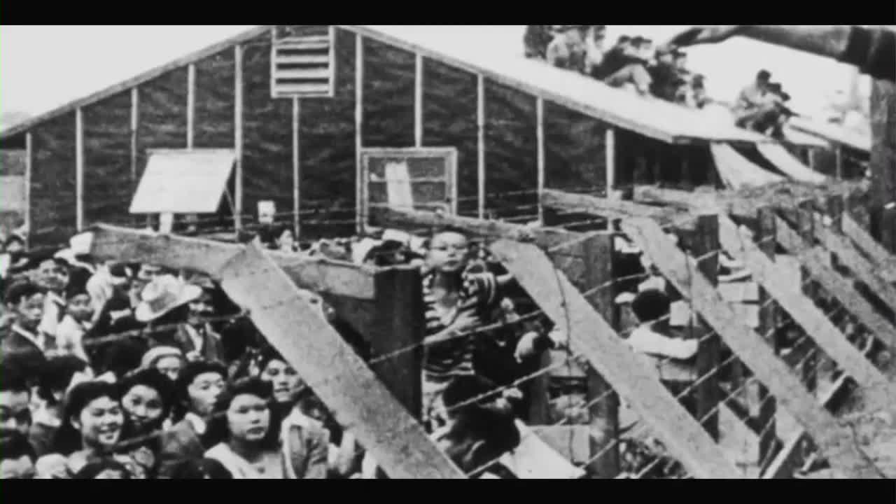 Behind a barbed-wire fence, a crowd of people, mostly women and children, are pressed together. Some sit on the roof of a barrack-like structure, watching.
