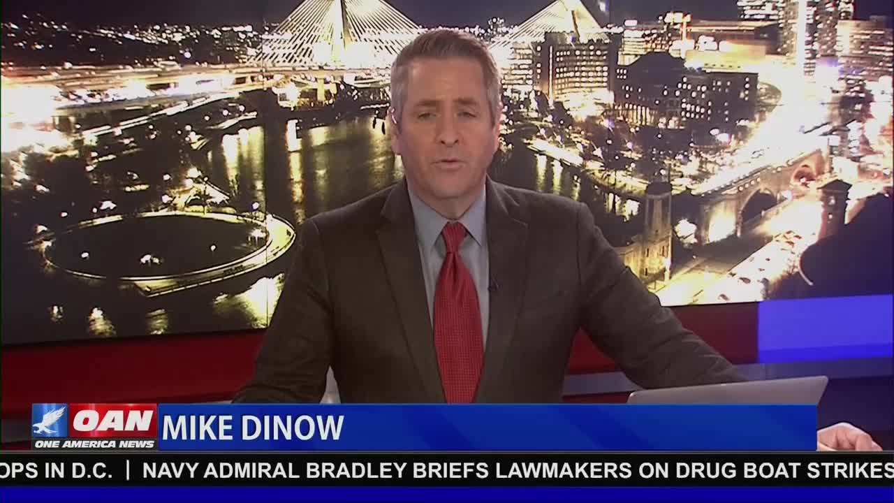 Mike Dinow, from One America News, is speaking from behind a desk. Behind him, a nighttime cityscape shimmers with lights.
