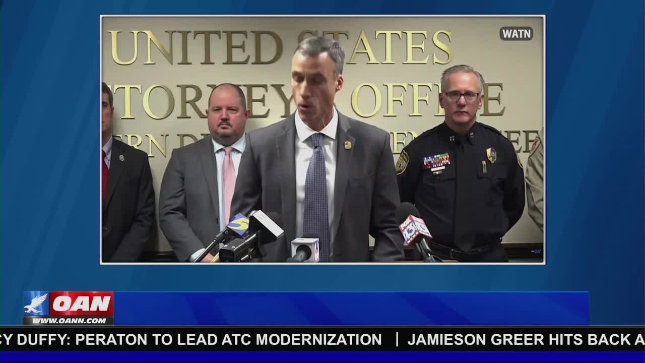 A man in a gray suit speaks into a cluster of microphones, flanked by other men in suits and a police uniform. The scene is broadcast on One America News Network, with a United States Attorney Office backdrop visible behind them.
