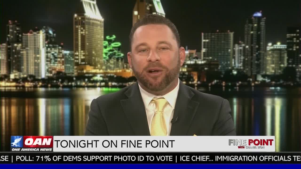 A man in a suit and yellow tie speaks directly to the camera. Behind him, the San Diego skyline glitters at night, reflected in the water.