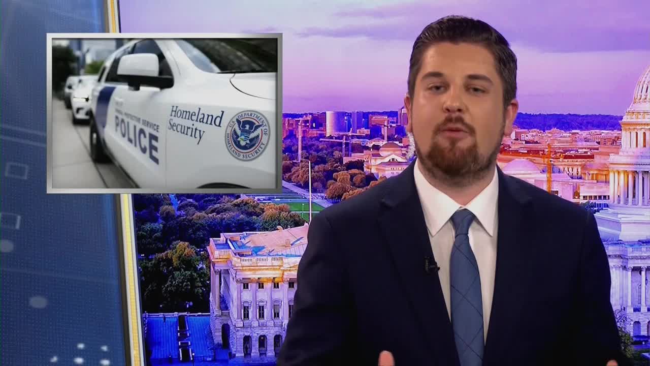A Homeland Security police vehicle sits parked, its white body emblazoned with official seals. A man in a suit speaks in front of a backdrop of the U.S. Capitol building.