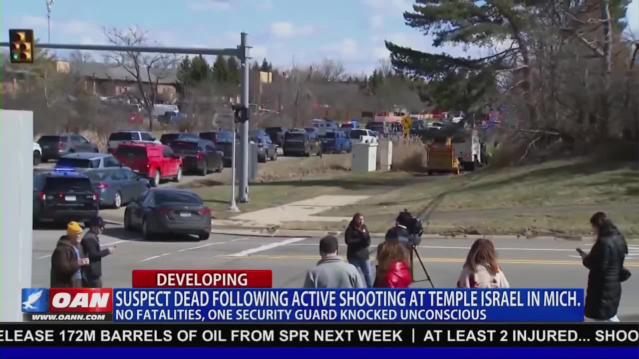 A line of police cars and other vehicles are parked along the road. A news crew is set up with a camera on a tripod, filming the scene.