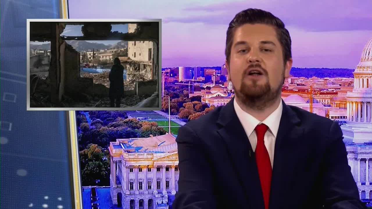 A man in a suit speaks directly to the camera, a red tie prominent against his dark jacket. Behind him, a cityscape unfolds, featuring the dome of the U.S. Capitol building. To his left, a small inset displays a somber scene of a person standing amidst the ruins of a building.