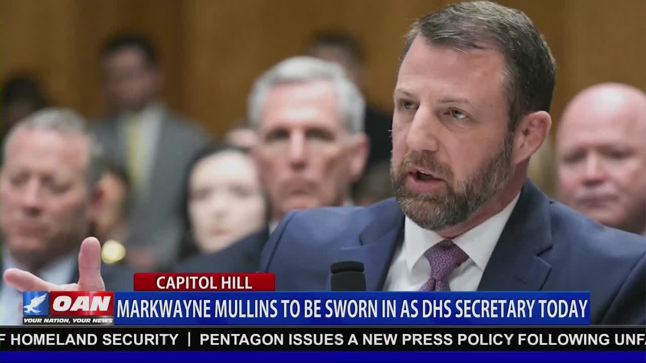 A man with a beard speaks into a microphone, his hand gesturing. Behind him, other men in suits listen intently in a room that looks like it's on Capitol Hill. A news chyron from OAN reads "MARKWAYNE MULLINS TO BE SWORN IN AS DHS SECRETARY TODAY."