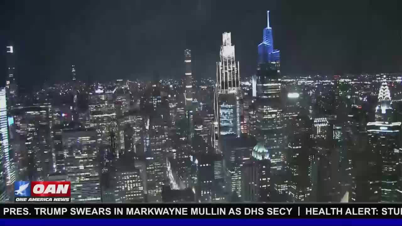 The New York City skyline glows with countless lights against the dark night sky. A banner at the bottom of the screen reads "PRES. TRUMP SWEARS IN MARKWAYNE MULLIN AS DHS SECY | HEALTH ALERT: STUD."
