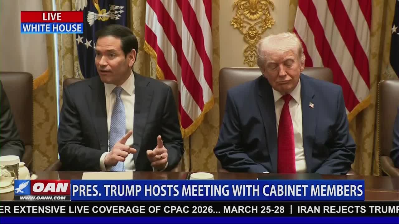 The man in the suit gestures with his hands as he speaks. Beside him, Donald Trump appears to be dozing with his eyes closed.