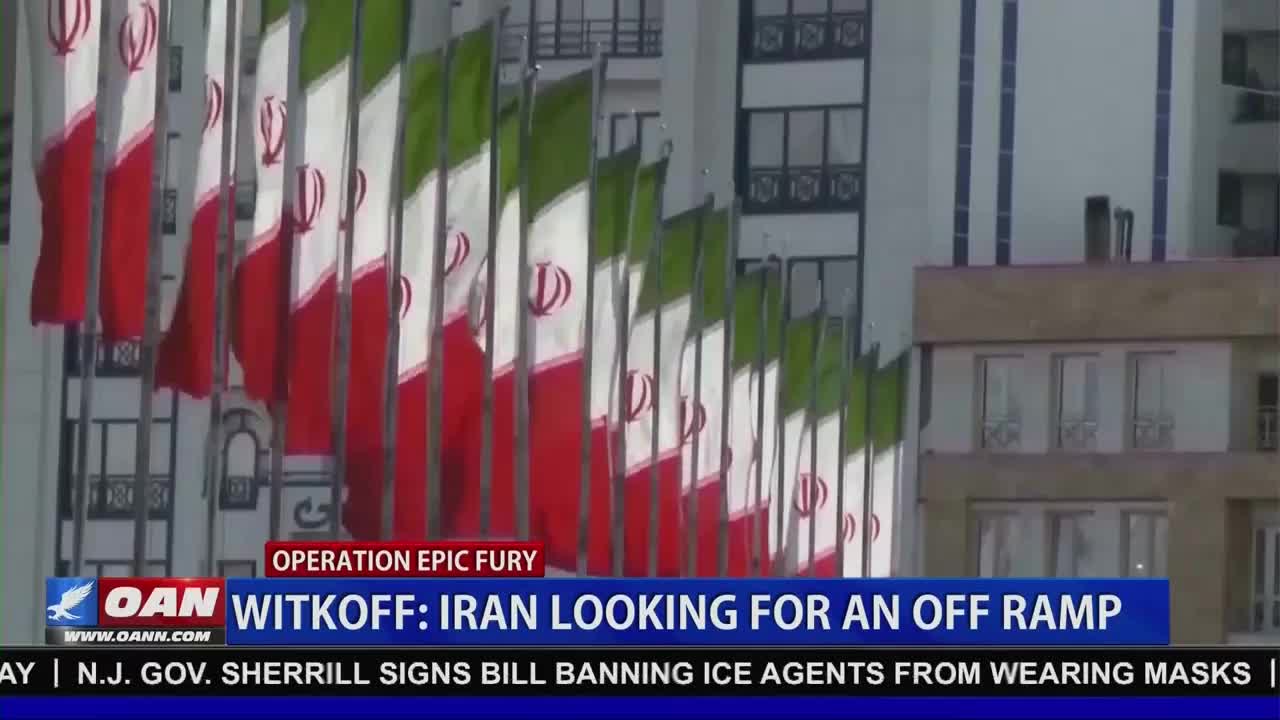 A row of Iranian flags ripples in the breeze against a modern building. A news chyron from One America News Network reads "WITKOFF: IRAN LOOKING FOR AN OFF RAMP."