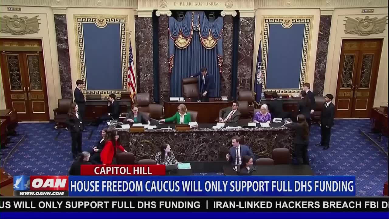 A man in a suit gestures from behind a large chair as others stand and sit at desks in a grand chamber. A banner scrolls across the bottom, announcing the House Freedom Caucus's stance on DHS funding.