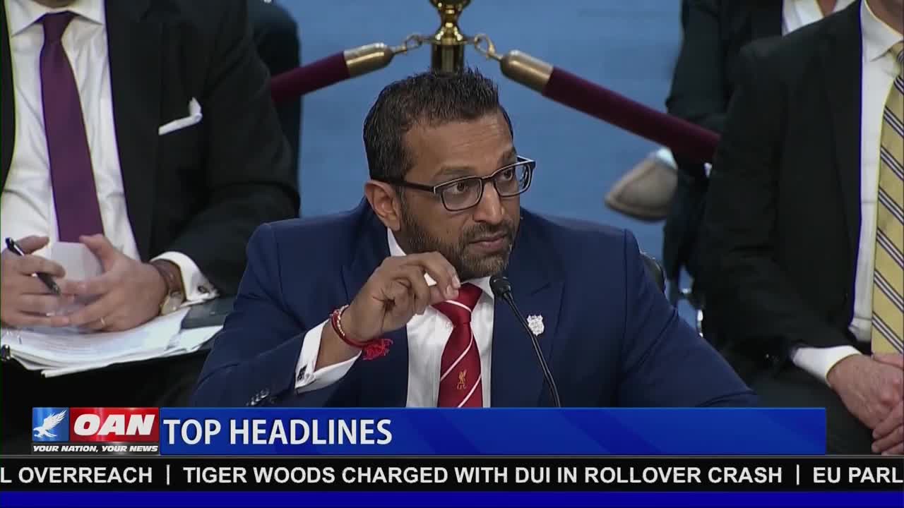 A man in a blue suit and red tie speaks into a microphone, gesturing with his right hand. The OAN chyron below displays "TOP HEADLINES" and mentions "TIGER WOODS CHARGED WITH DUI."
