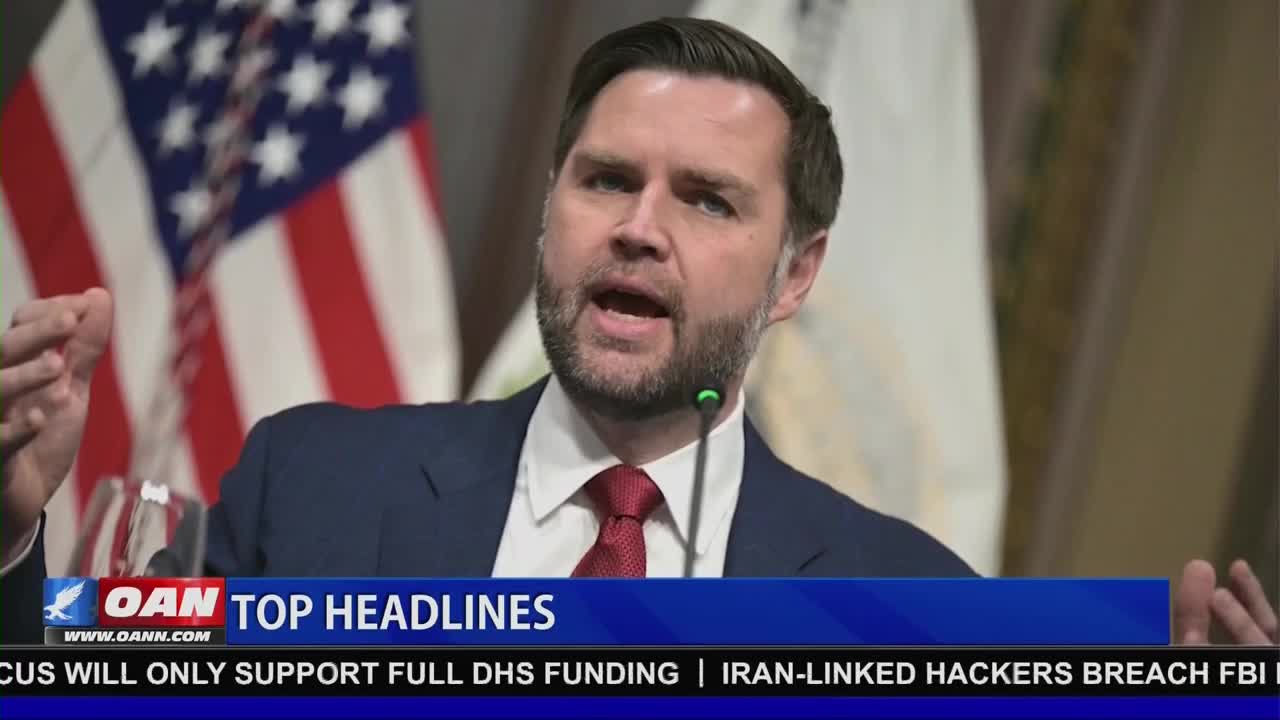 A man in a dark suit and red tie speaks at a podium, his right hand gesturing upward. Behind him, the American flag hangs, slightly out of focus.