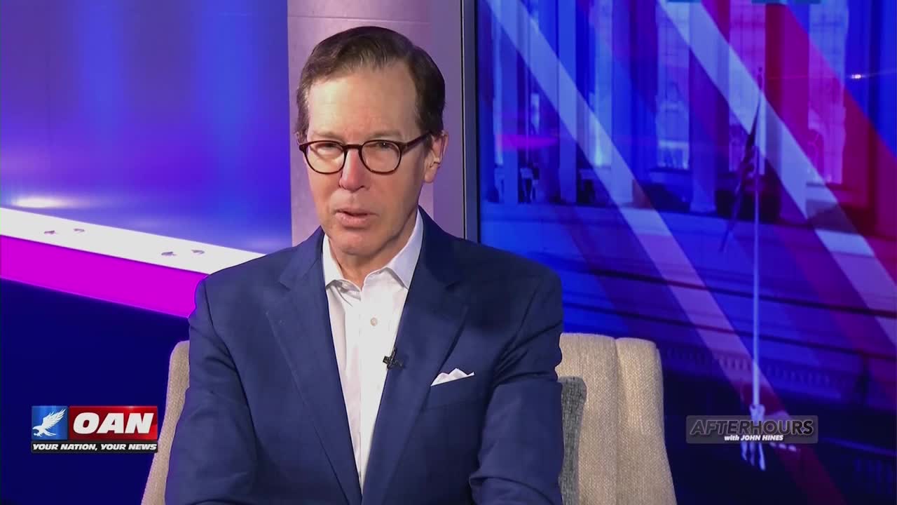 A man in a blue suit and glasses sits on a couch, speaking into a microphone. Behind him, a screen displays the One America News Network logo and a graphic of a building.