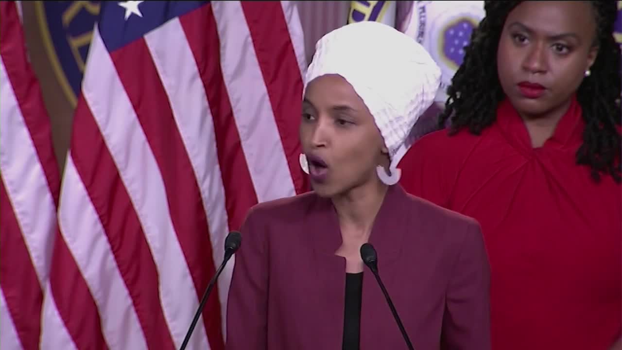 A woman in a white headscarf speaks into a microphone, her mouth open in emphasis. Behind her, American flags hang next to another woman in a bright red top.
