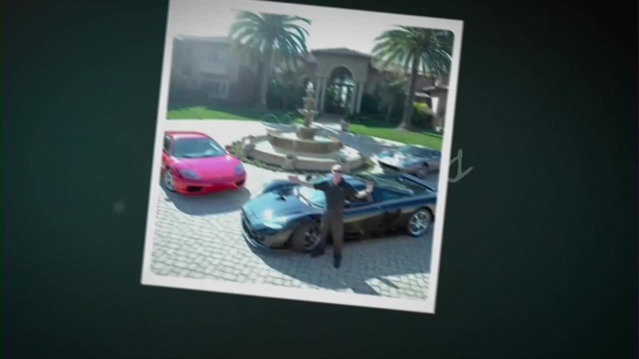 A man stands with his arms outstretched in front of a sleek, dark sports car. Behind him, a bright pink Ferrari and another silver car are parked on a cobblestone driveway.