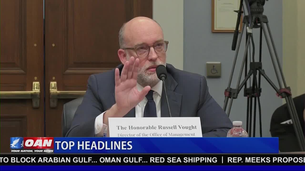 Russell Vought, Director of the Office of Management, raises his hand as he speaks at a hearing. A One America News Network chyron displays "TOP HEADLINES" below him.
